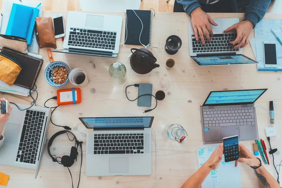 Overhead view of a shared work table — multiple laptops, phones, notebooks, and headphones spread across it